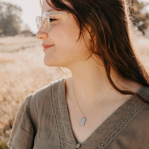 A young woman with long brown hair and clear glasses smiles outdoors in a sunlit field, wearing a textured olive green top and the Long Live Bar Necklace, a sterling silver religious accessory from Viva Cristo Rey jewelry.