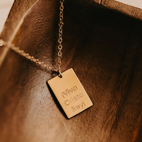 The Long Live Bar Necklace, a 14k gold filled rectangular pendant engraved with ¡Viva Cristo Rey!, is displayed in a wooden dish—an ideal piece for anyone seeking meaningful Viva Cristo Rey jewelry or a special charm.