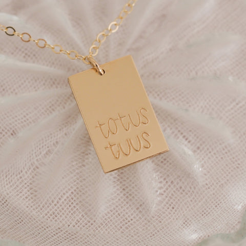 The Totus Tuus Necklace showcases a 14k gold filled rectangular pendant engraved with totus tuus on a delicate chain, symbolizing devotion to Mary. It is displayed beautifully on a textured white fabric background.
