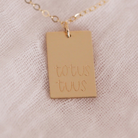 The Totus Tuus Necklace, crafted in 14k gold fill with a rectangular “totus tuus” pendant symbolizing Marian devotion, is elegantly displayed on textured white fabric.