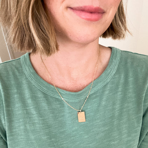 A woman with short, light brown hair wears a green top and a Totus Tuus Necklace. The 14k gold filled rectangular pendant features delicate engraving, symbolizing her devotion to Mary. The photo is cropped to show only her neck and shoulders.
