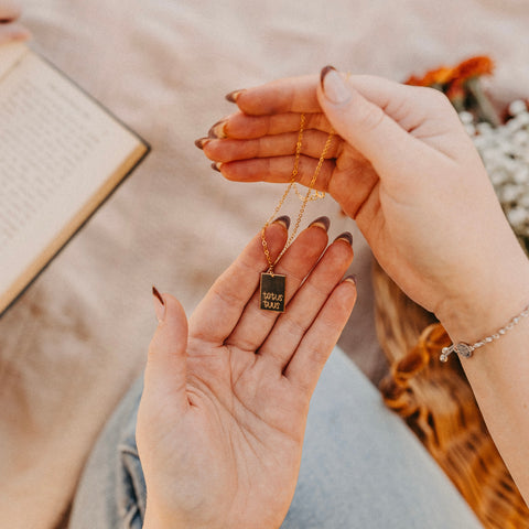 A person holds the Totus Tuus Necklace—14k gold filled, featuring a dark pendant inscribed with Totus Tuus in elegant script, symbolizing devotion to Mary. Their manicured nails stand out against a blurred book, basket, and flowers in the background.