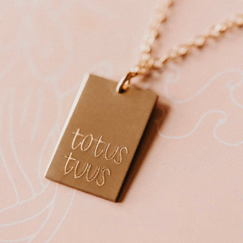 The Totus Tuus Necklace is a 14k gold filled piece with a rectangular pendant engraved totus tuus, symbolizing devotion to Mary, displayed on a light pink surface.