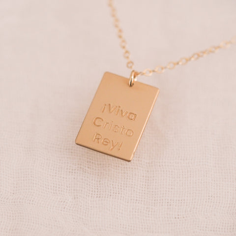 The Long Live Bar Necklace, a 14k gold filled bar engraved with “¡Viva Cristo Rey!” on a delicate chain, shines beautifully against a light textured fabric background.