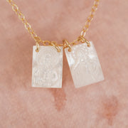 The Scapular Style Necklace features a 14k gold-filled chain and two rectangular Mother of Pearl charms engraved with religious figures, displayed on a soft peach-colored background.